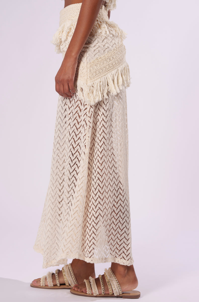 A side view of a person wearing the ON THE FRINGE SKIRT, a beige, boho-chic crochet-patterned fringe skirt featuring intricate detailing at the waist. The outfit is perfectly paired with beige embellished flat sandals, making it an ideal addition to a festival wardrobe. The background is plain white.