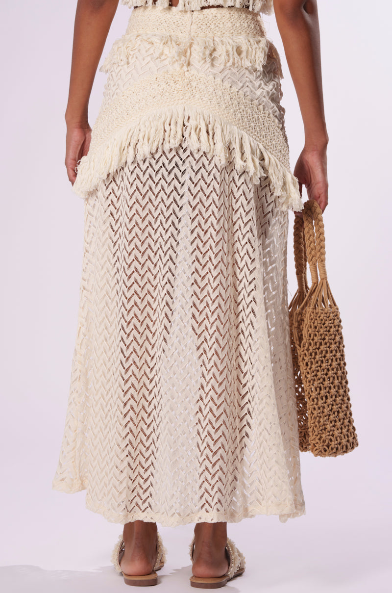 A person is wearing the ON THE FRINGE SKIRT, a cream-colored crochet mesh piece with fringed details. The skirt showcases a chevron design, making it an ideal addition to any festival wardrobe. They are holding a woven straw bag and standing against a plain background.