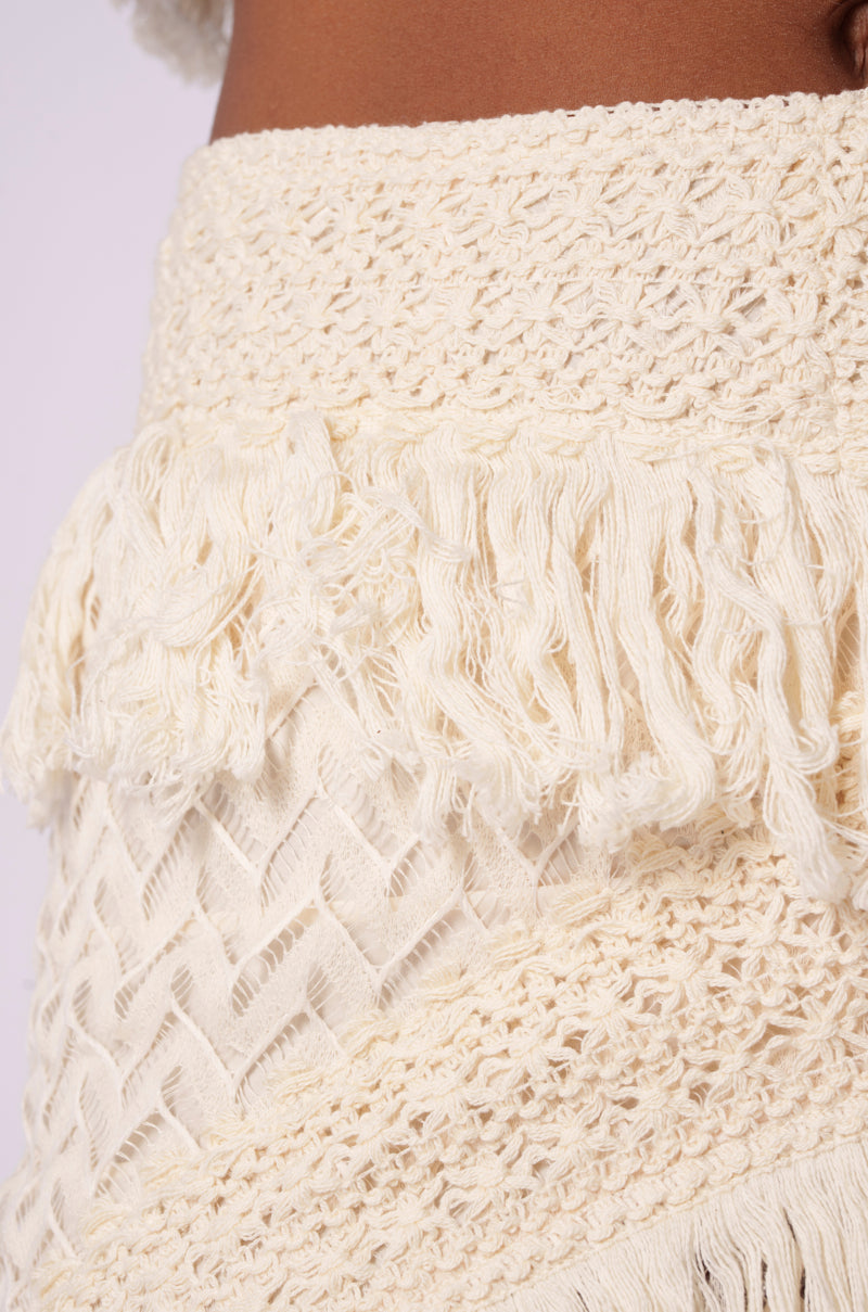 Zoomed-in view of the cream-colored ON THE FRINGE SKIRT, showcasing the intricate knit patterns and fringe details. The texture displays layered, woven sections adorned with diamond shapes and a band of loose, hanging threads. Ideal for your festival wardrobe, this bohemian-style skirt offers a hand-crafted appearance.