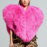 Confidently styled, a person showcases the STAR CROSSED LOVER FAUX FUR HEART VEST, a large and fluffy pink heart-shaped piece, layered over a bold leopard print ensemble. With their hair braided, they pose against a simple gray background.