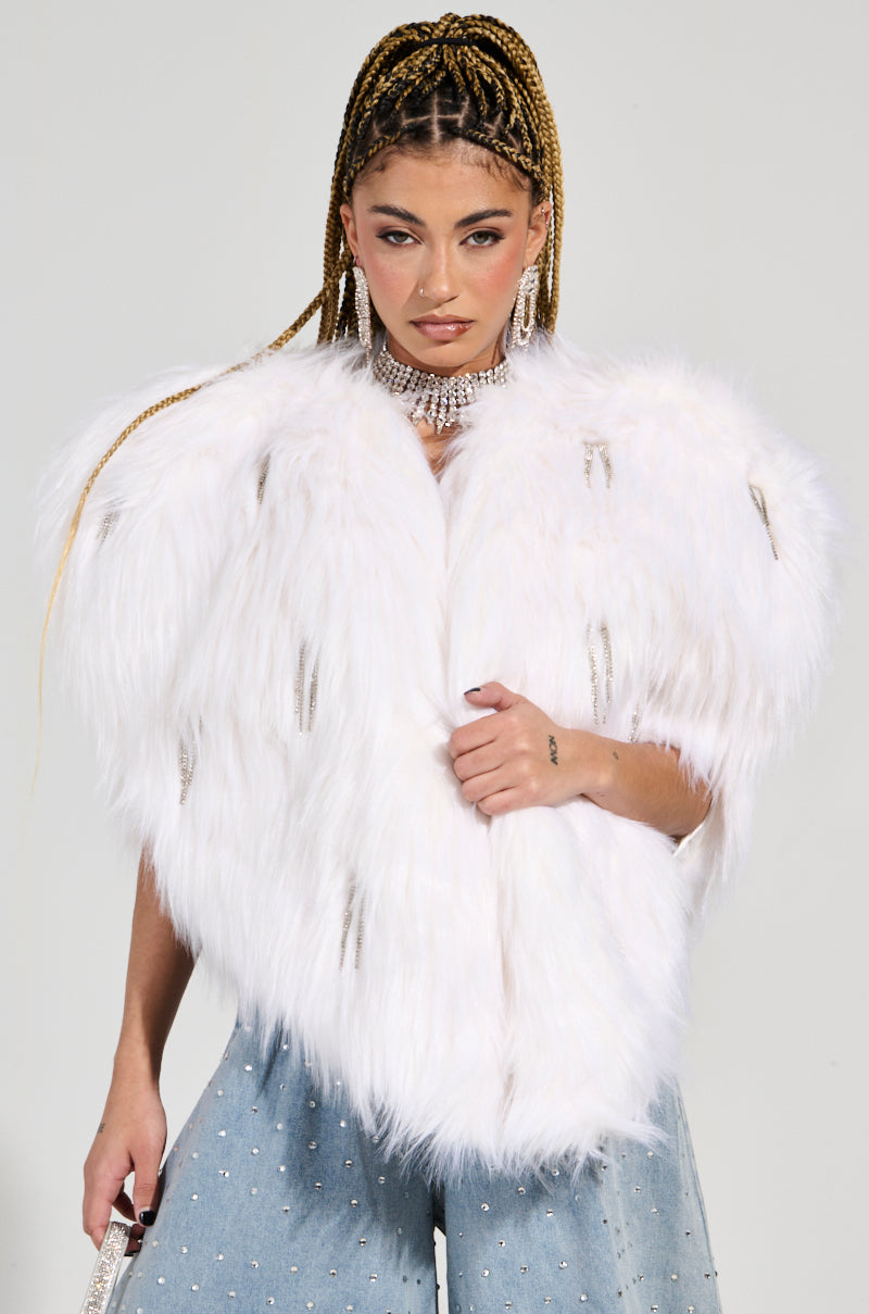 A person with braided hair is wearing a glamorous LIMITED EDITION STAR LOVER FAUX FUR HEART VEST IN WHITE and blue jeans adorned with rhinestones. They are accessorized with a sparkling choker necklace and earrings, standing against a plain background.