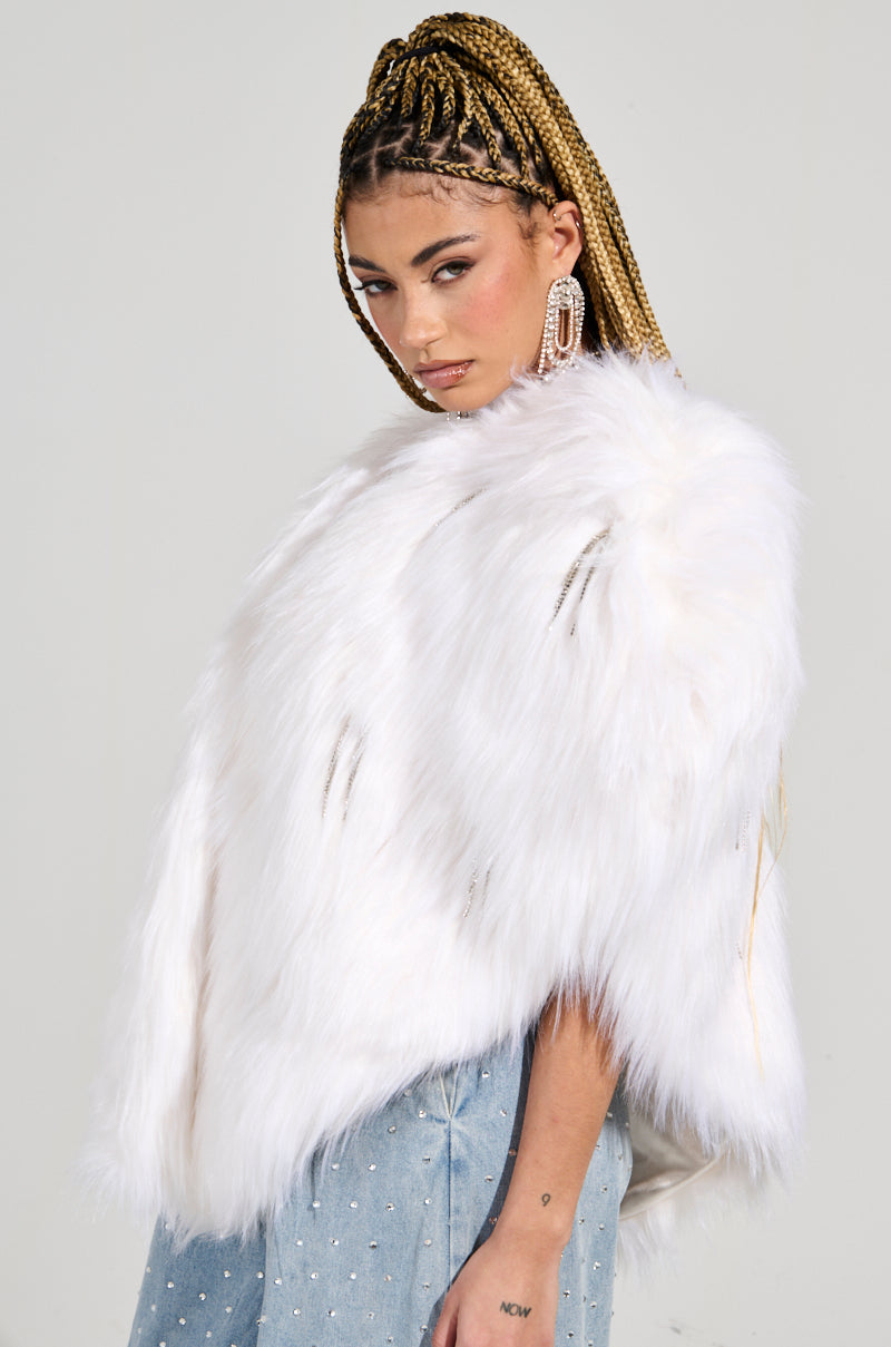 A person with braided hair poses confidently, wearing the LIMITED EDITION STAR LOVER Faux Fur Heart Vest in White and large, sparkly earrings. The ensemble is completed by an AKIRA Label light blue skirt adorned with small shiny accents against a plain backdrop.