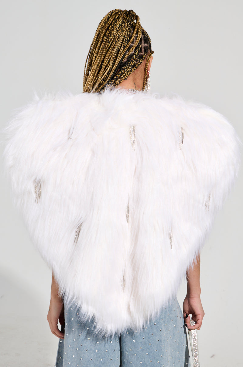 A person with braided hair stands facing away, wearing a luxurious statement piece—a Limited Edition Star Lover Faux Fur Heart Vest in white adorned with decorative elements. Complementing the outfit are light blue jeans with rhinestone embellishments, and they hold a small purse. The background is plain gray.