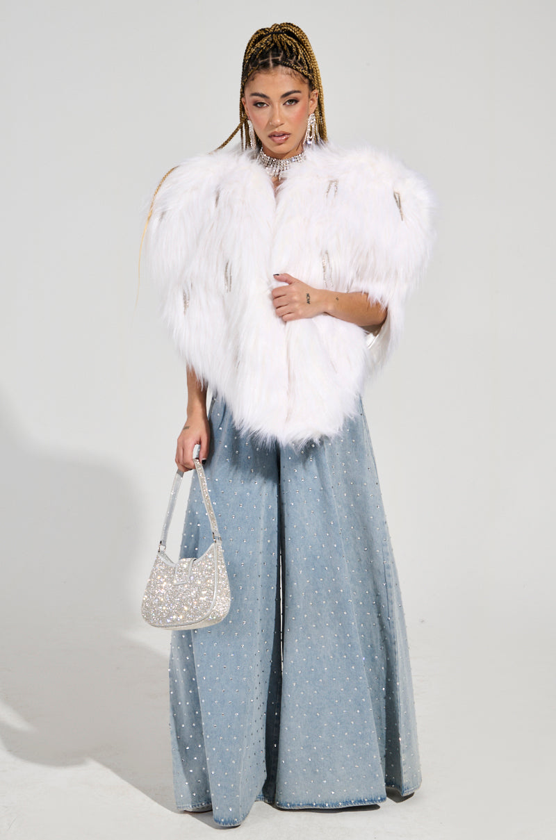 Wearing an exquisite LIMITED EDITION STAR LOVER FAUX FUR HEART VEST IN WHITE paired with wide-legged blue pants adorned with rhinestones, the individual holds a shimmering silver handbag. Their long braided hair adds an elegant touch as they pose against a simple background.