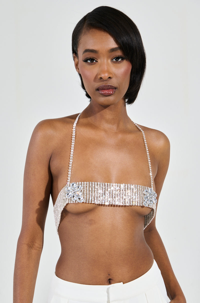 Against a plain backdrop, a woman with short, dark hair exudes confidence in her sparkling bejeweled top and white pants. The STAR GURL BLING BODY CHAIN adds an extra layer of elegance as she stands with one hand on her hip.