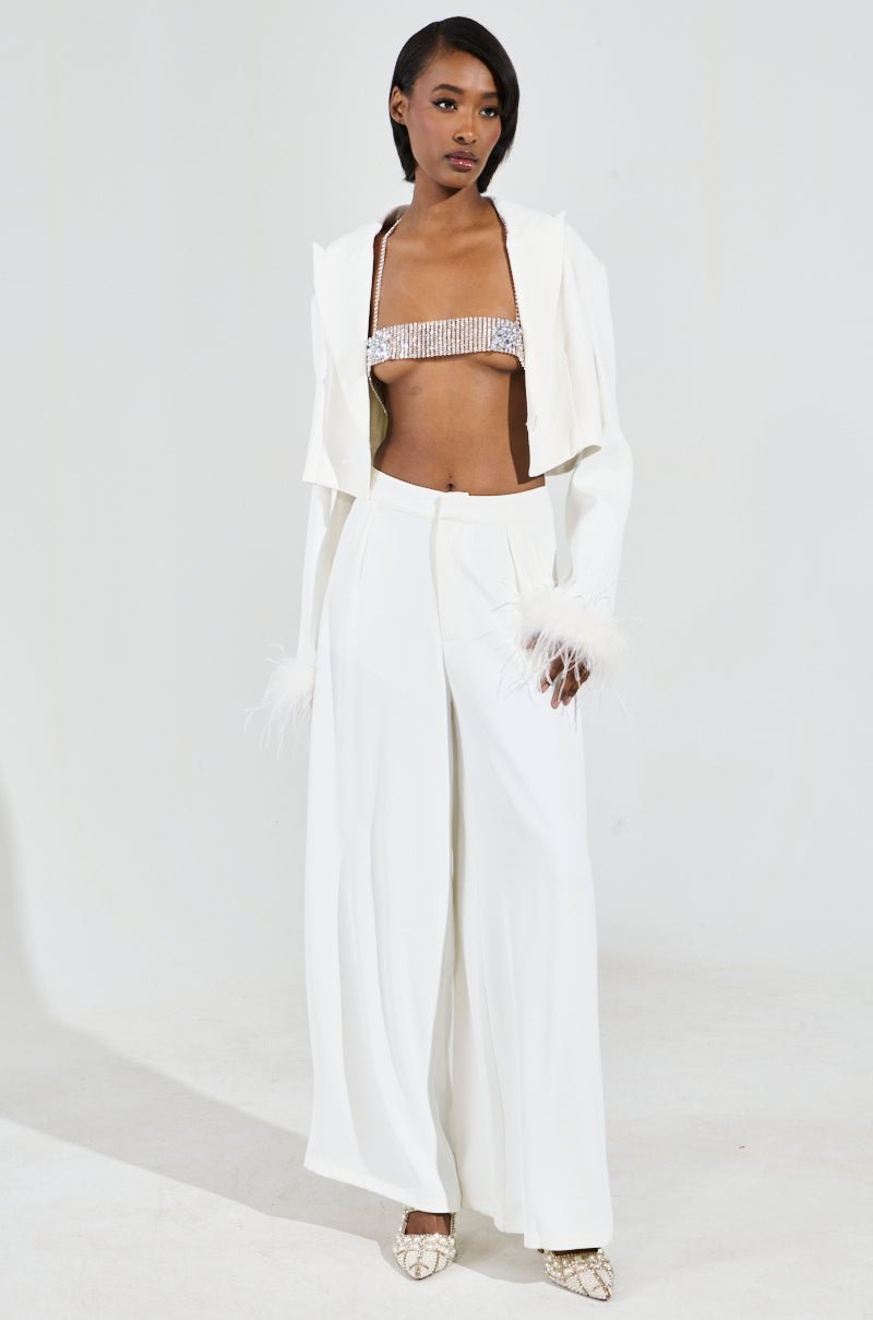 The woman dazzles in a fashionable white outfit featuring a cropped jacket with feathered cuffs, a beaded top, and wide-legged pants. She enhances her look with the STAR GURL BLING BODY CHAIN for extra shimmer, along with silver jewels and pointed shoes, standing confidently against a plain backdrop.