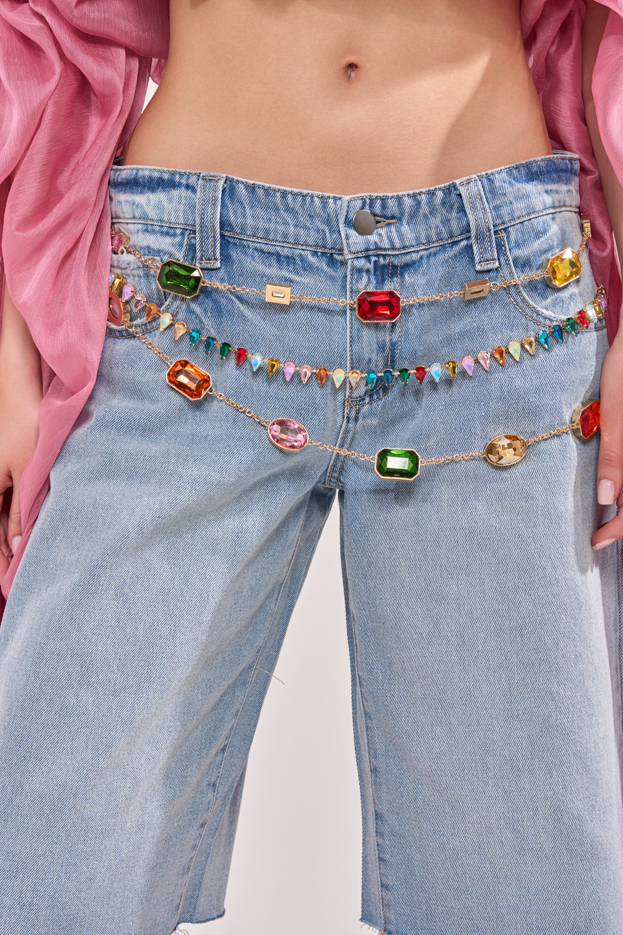 A person wears light blue jeans accessorized with the BELIEVE IT CHAIN BELT draped across the waistband, paired with a pink flowing top. The image is cropped to show only the lower torso and upper legs.