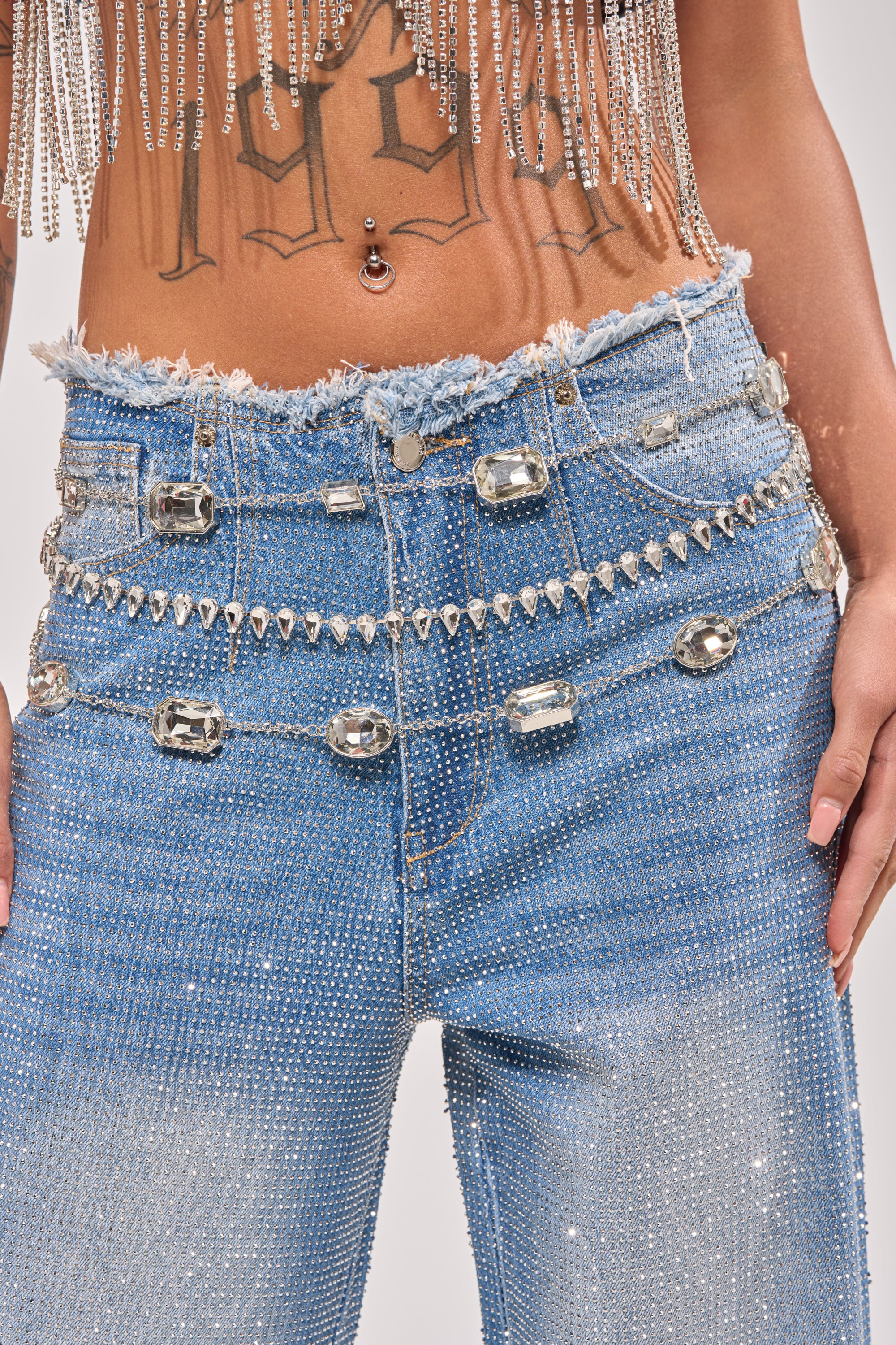 A person wears light blue rhinestone-studded jeans with a frayed waistband, accessorized with the GLITZ AND GLAM CHAIN BELT. Their bare midriff features a belly button piercing and a "1996" tattoo. 0