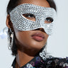 A person is adorned with the MASQUERADE MASK, which is sparkling silver and embellished with gemstones. They pair it with large star earrings and a sequined outfit showcasing floral patterns, all set against a soft, neutral background.