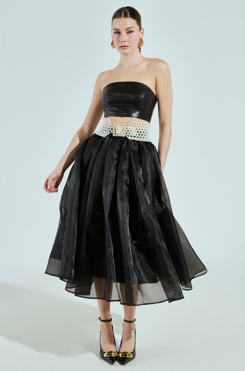A woman models a strapless black dress with a full skirt and adjustable fit, accentuated by the PEARL PLEASE BELT. She accessorizes with gold hoop earrings and black ankle-strap heels featuring gold pointed toes, posing elegantly against a plain light background.