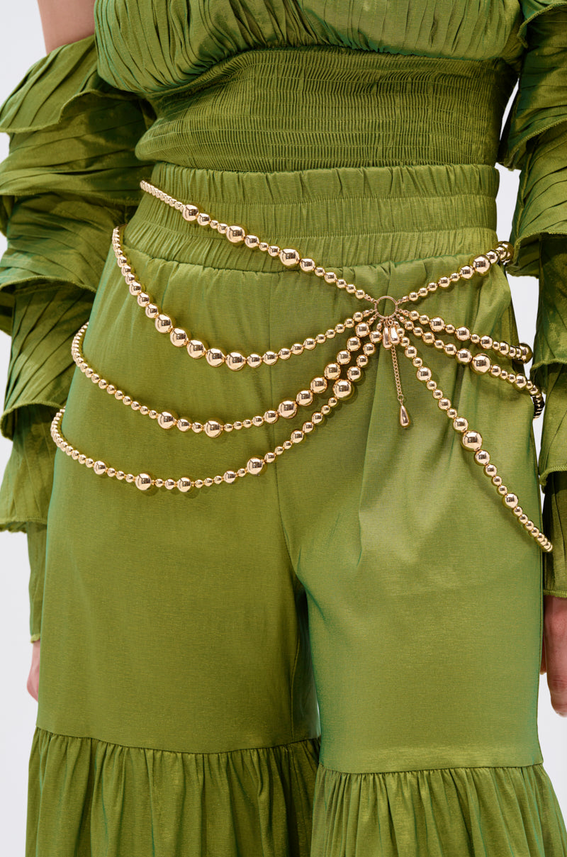 A person in a shiny, ruched green outfit accessorized with the GOLDEN HOUR BEAD BELT, a sculptural, multi-layered gold-tone waist belt draped elegantly around their waist and hips.