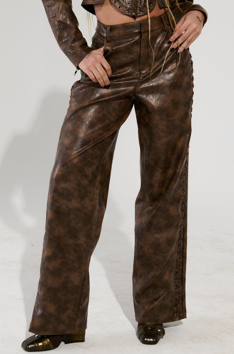 The individual sports the chic UP TO IT LACE UP TROUSERS, which are high-waisted and made of faux leather, featuring intricate lace-up details along the sides. These pants have a marbled texture that stands out against the light background and are paired with a matching AKIRA Label jacket.