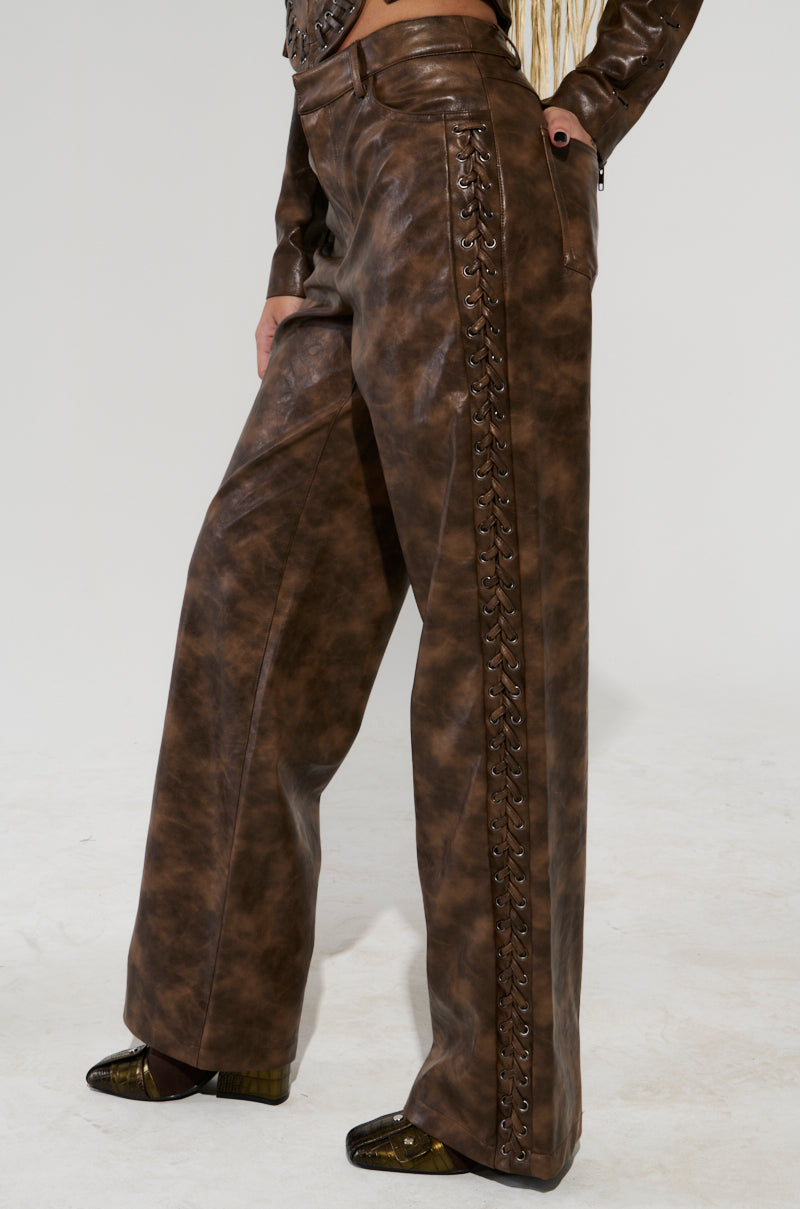 A person is clad in the UP TO IT LACE UP TROUSERS, featuring brown faux leather and intricate lace-up detailing along the sides. Paired with dark shoes and a glimpse of a matching leather jacket, they stand against a plain background.