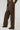 A person is clad in the UP TO IT LACE UP TROUSERS, featuring brown faux leather and intricate lace-up detailing along the sides. Paired with dark shoes and a glimpse of a matching leather jacket, they stand against a plain background.