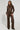 A person with long, braided hair is dressed in an AKIRA Label brown faux leather outfit featuring the UP TO IT LACE UP TROUSERS. The matching fitted jacket is adorned with decorative details as they stand against a plain white background, looking confidently at the camera.