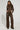 A person with braided hair poses confidently in an AKIRA Label two-piece outfit, sporting a textured brown long-sleeve top and the UP TO IT LACE UP TROUSERS. The ensemble, crafted from faux leather, features intricate details and stitching. The background is plain white.
