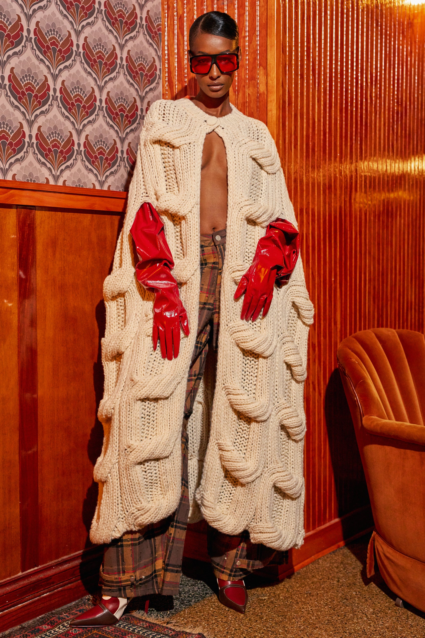 A person wears a cream-colored TEXTURED CHUNKY KNIT SWEATER in a longline, open-front design, styled with bright red glossy gloves, red-tinted sunglasses, and plaid trousers.
