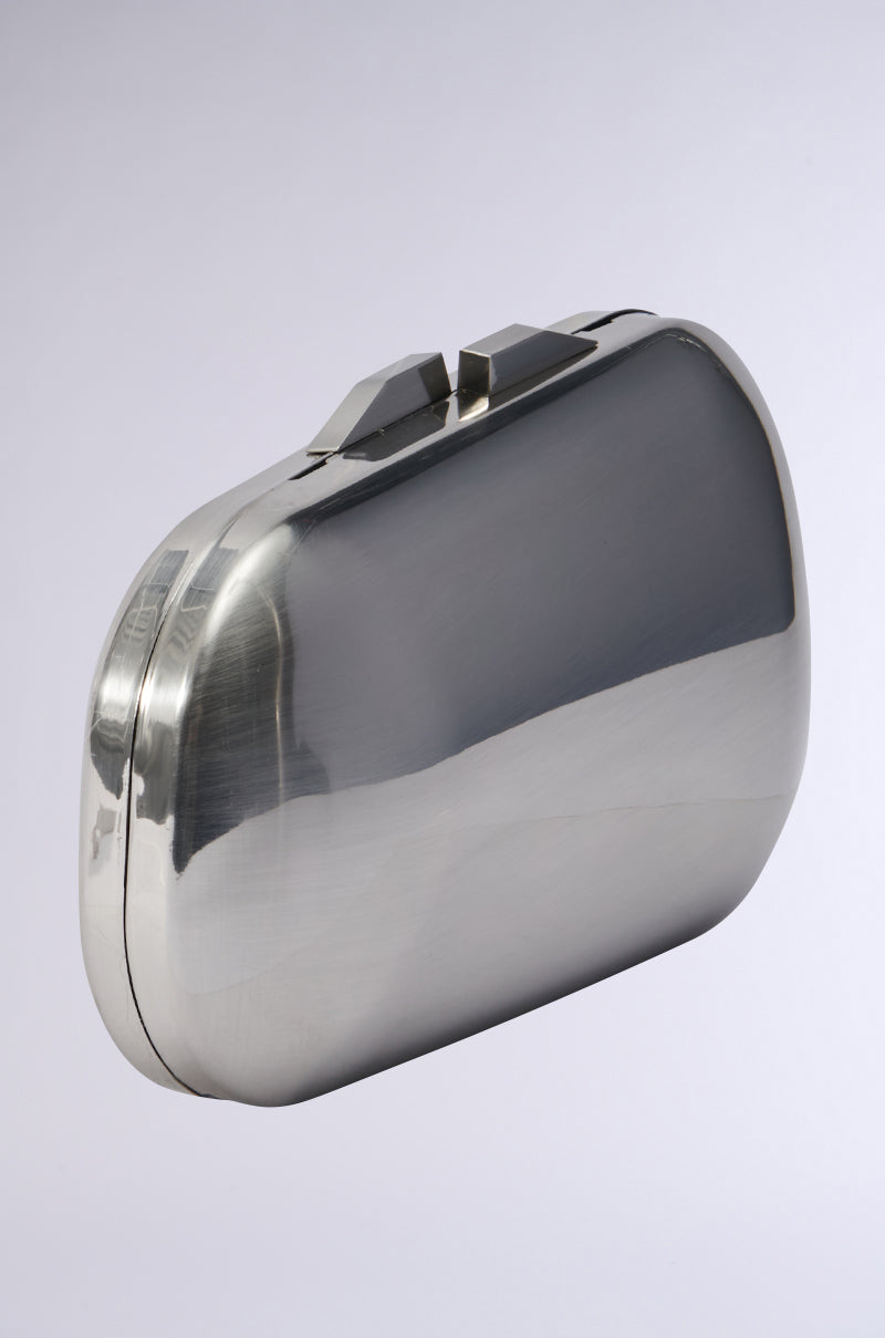 The METAL MANIA CLUTCH is a stylish, minimalist accessory featuring a rounded rectangular design. Its metallic silver finish elegantly reflects light, and the top is accented with a simple clasp. Presented against a plain light gray background, it comes with an optional detachable chain link strap for added versatility. 2