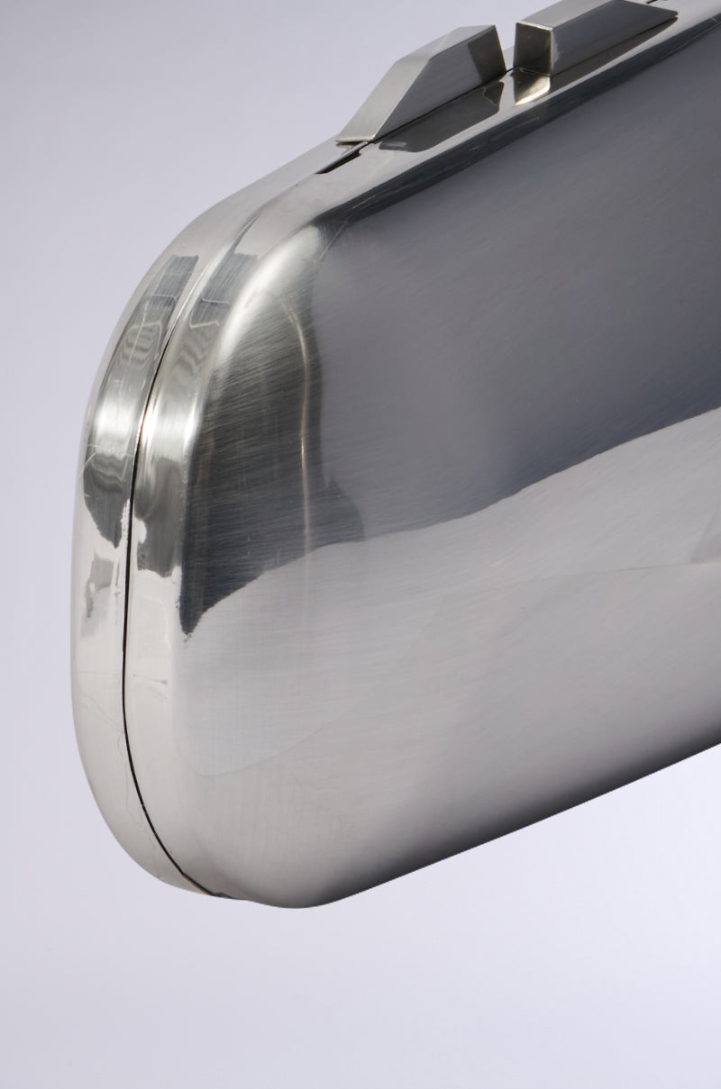 A close-up view of the METAL MANIA CLUTCH reveals its shiny, curved design and reflective metallic surface. The plain light-gray backdrop highlights its polished texture, and the detachable chain link strap contributes an elegant touch. 3