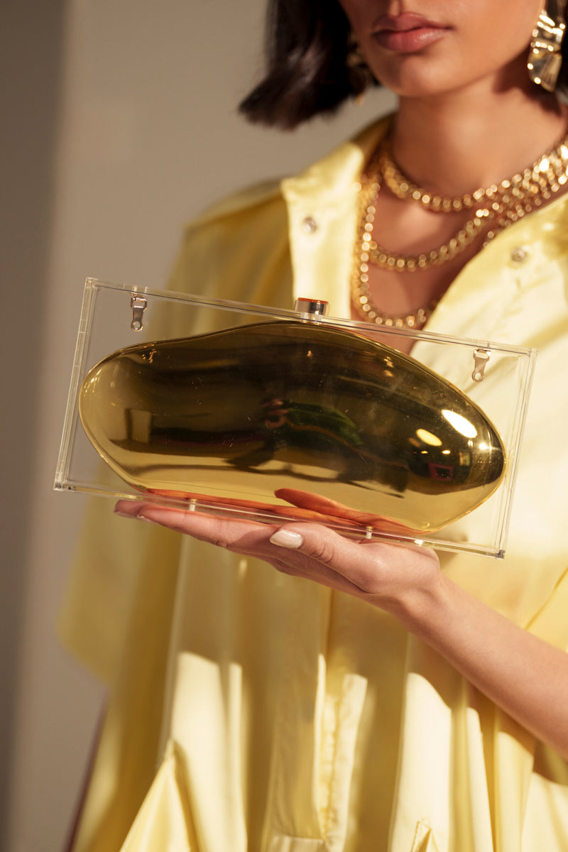 A woman in a yellow satin outfit holds the GOLDEN GODDESS CLUTCH, an acrylic bag featuring a shiny gold abstract shape. She accessorizes with layered gold necklaces, earrings, and has neatly manicured nails.