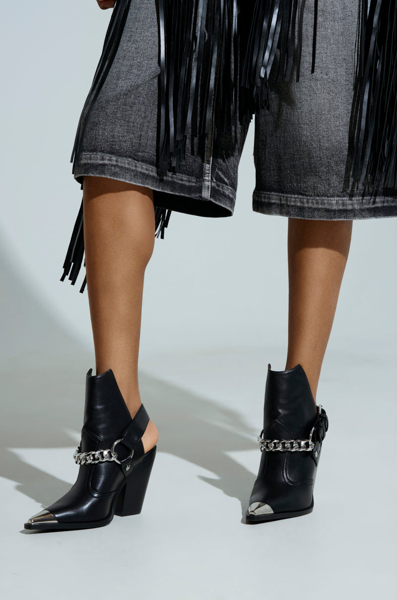 Someone is wearing fringed, knee-length denim shorts paired with the AZALEA WANG WADE BLACK WESTERN BOOTIE, featuring silver chains and pointed toes. A plain, light-colored background highlights the edgy ensemble.
