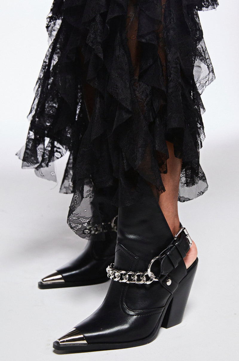 Close-up of someone wearing AZALEA WANG WADE black Western booties, featuring pointed toes, silver chain detailing, metallic tips, chunky heels; paired with a long black lace skirt with ruffled layers. Vegan leather completes the stylish ensemble.