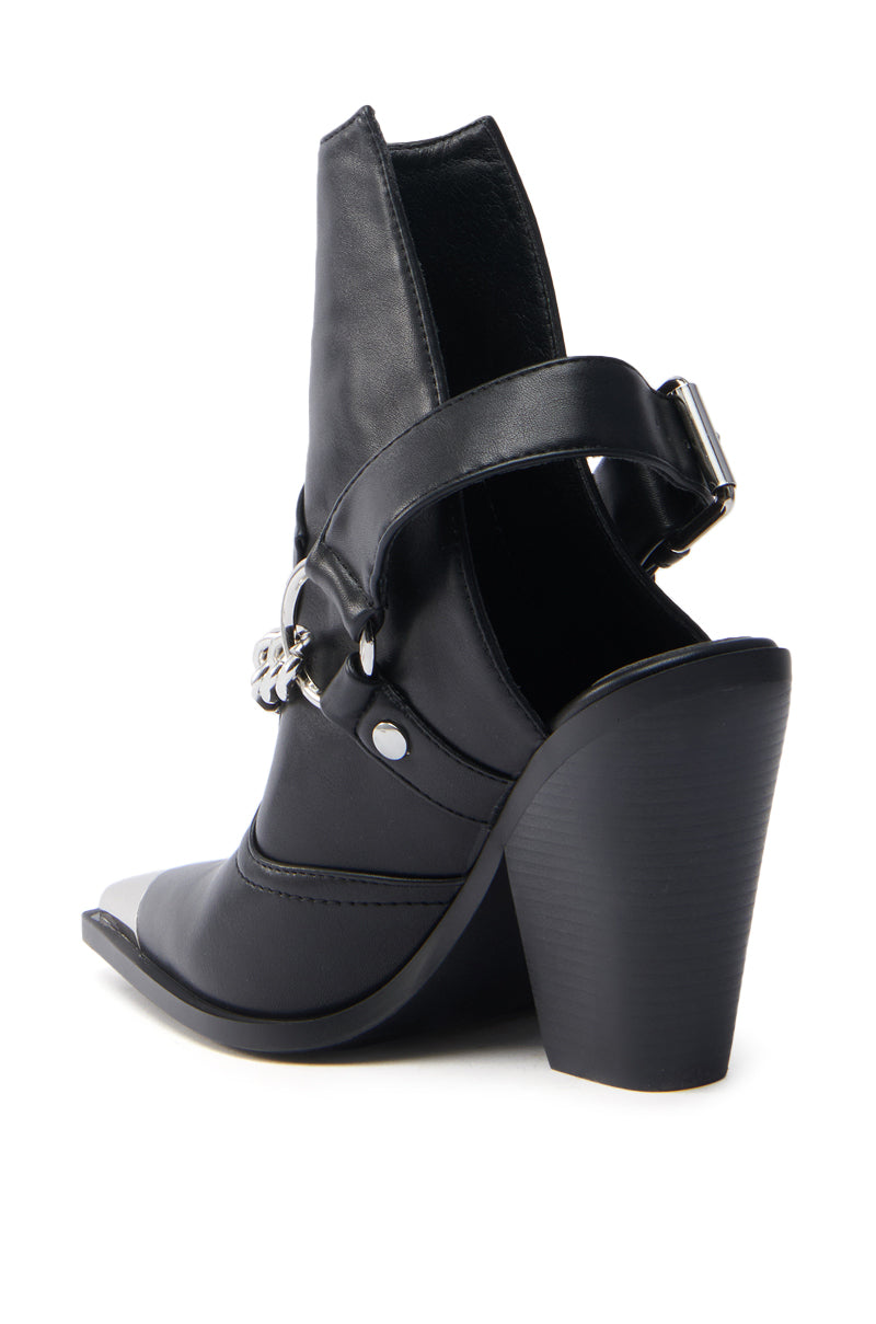 The AZALEA WANG WADE BLACK WESTERN BOOTIE is a high-heeled, black vegan leather shoe with an open back, pointed toe, silver chain detail, and a thick buckle strap around the ankle. Its chunky angled heel adds a modern twist to the Western bootie-inspired design.