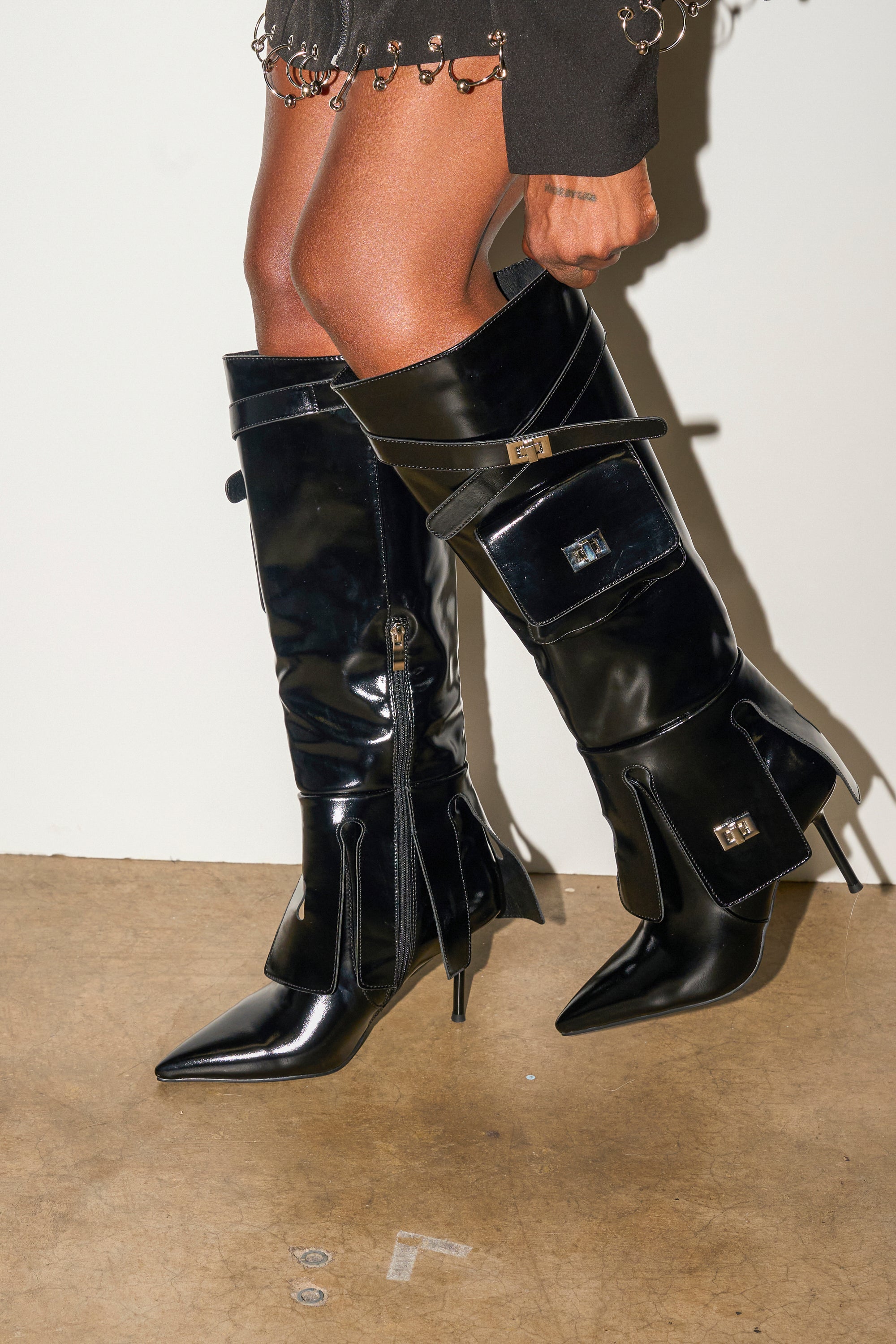 A person wears the AZALEA WANG WATERS BLACK STILETO BOOT WITH POCKETS, featuring shiny black material, pointed toes, buckles, and pocket details. The hem of a skirt with metallic rings is partially visible above the boots.