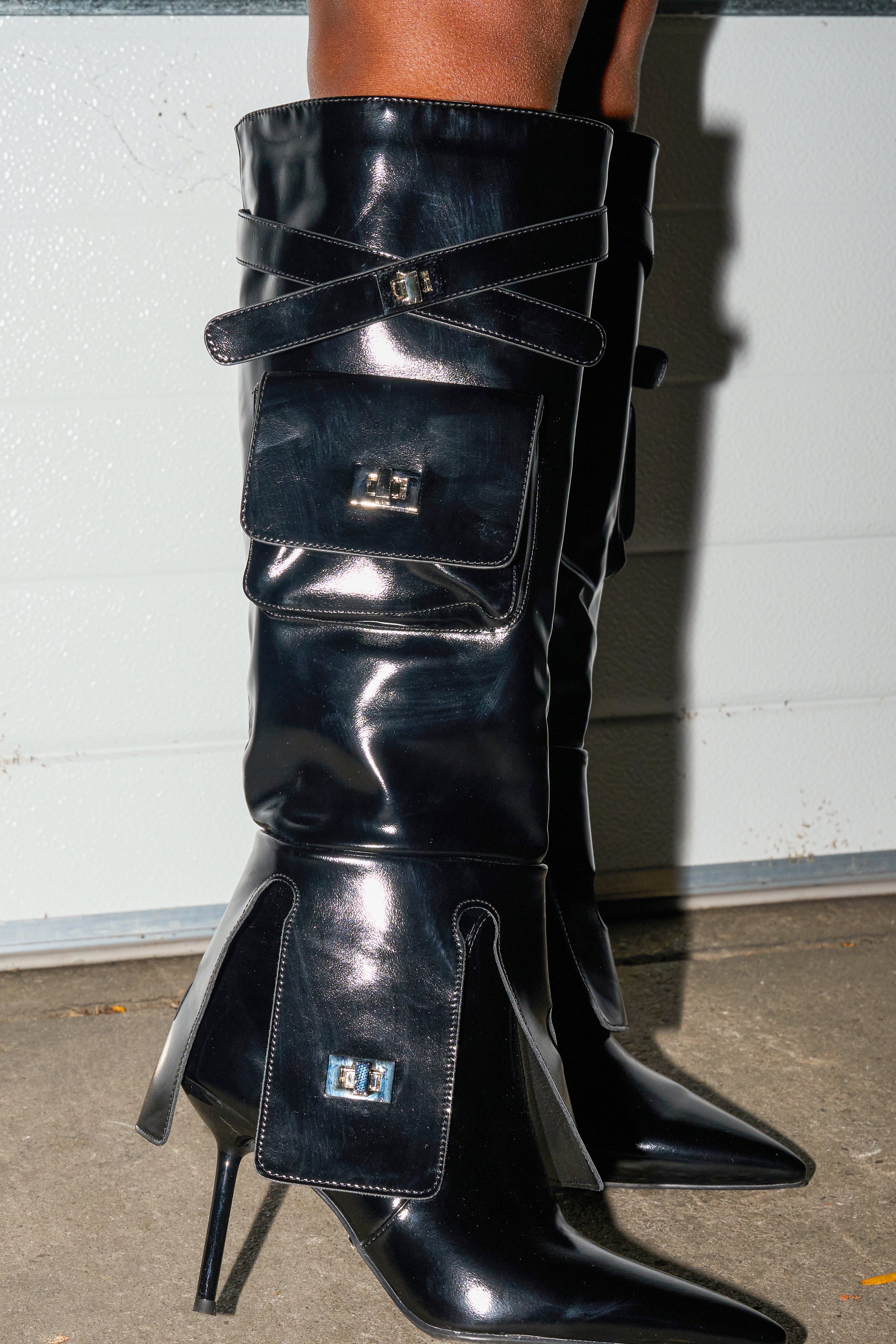 A close-up of a person wearing AZALEA WANG WATERS BLACK STILETTO BOOTS WITH POCKETS, featuring shiny black faux leather, pointed toes, stiletto heels, and buckle-secured decorative pockets against a light wall and concrete ground.