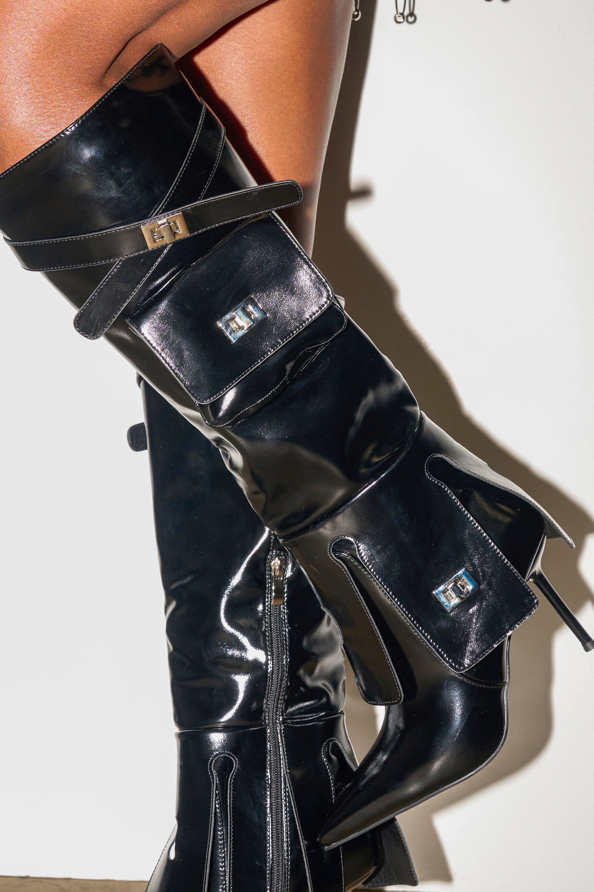 Close-up of a person wearing the AZALEA WANG WATERS BLACK STILETTO BOOT WITH POCKETS, featuring shiny black pointed toes, attached side pockets with metallic clasps, plus decorative straps and zippers for extra style.