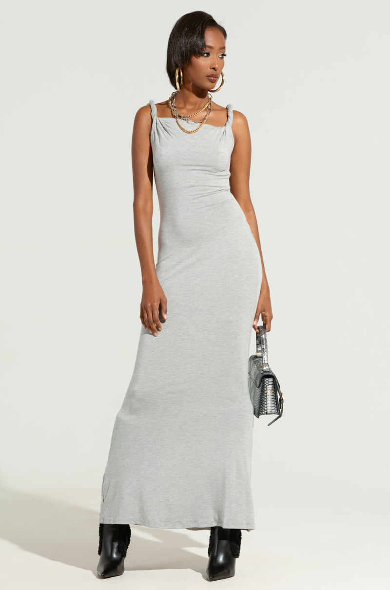 A woman wearing THE MAIN EVENT TWISTED MAXI DRESS, a sleeveless gray maxi dress with a bodycon fit and asymmetrical neckline, stands against a plain background. She accessorizes with a necklace, hoop earrings, and black ankle boots. Holding a small handbag, her hair is styled straight.