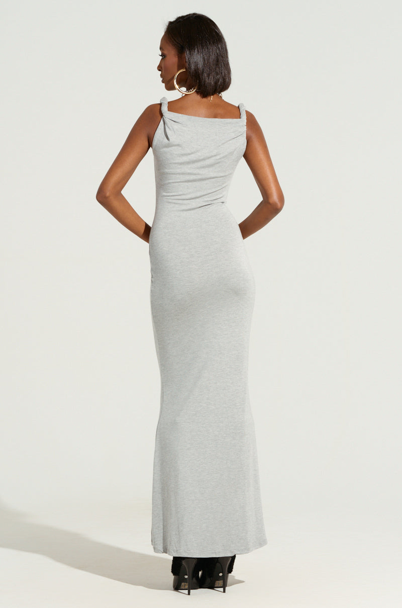 A woman stands facing away, adorned in the long, form-fitting THE MAIN EVENT TWISTED MAXI DRESS that features an asymmetrical neckline. With short hair flowing freely, she accessorizes with large hoop earrings and completes her look with a pair of black heels.