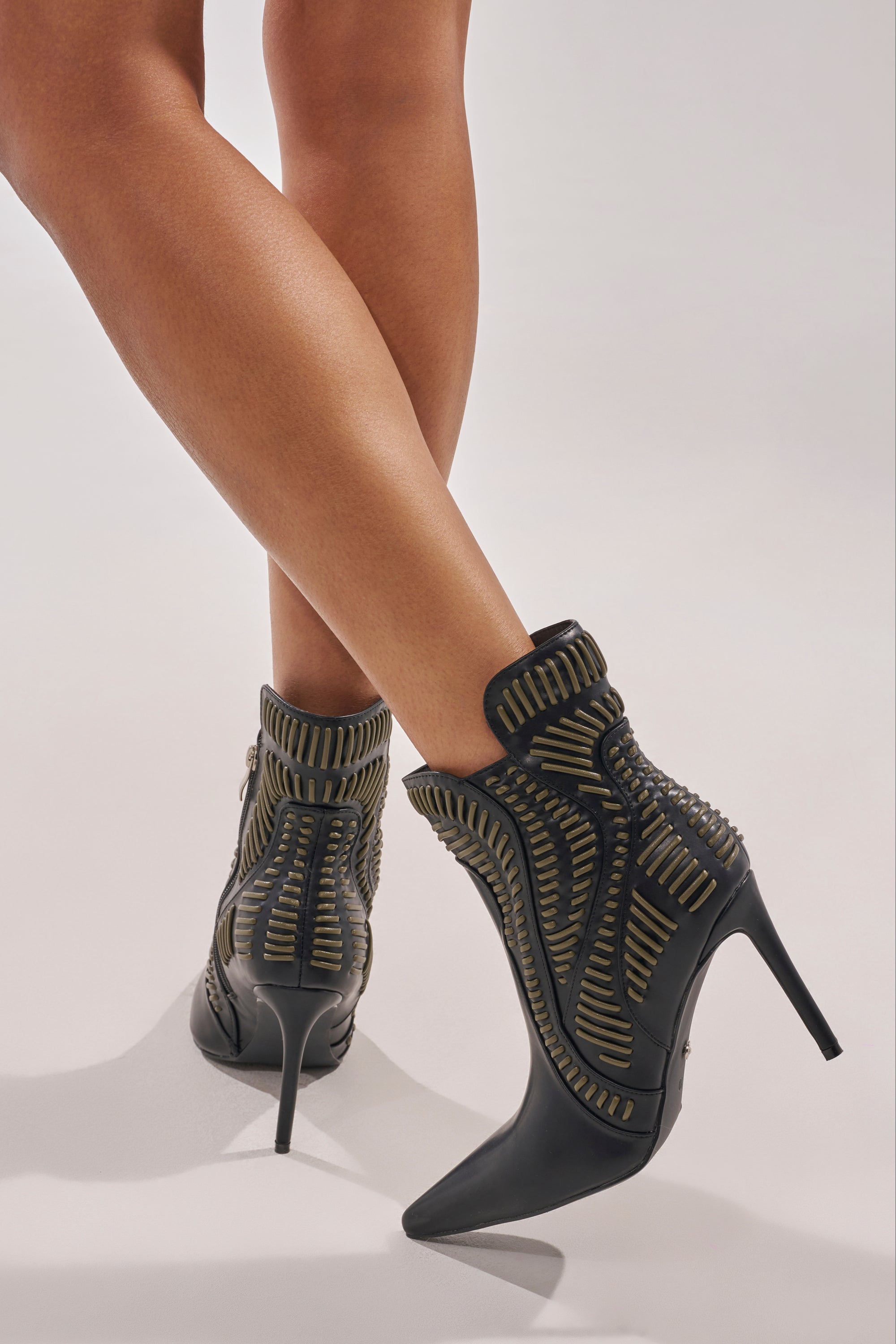 Someone stands with legs crossed on a light background, wearing the AKIRA GOLD WEAVER BLACK AND OLIVE BOOTIE—black ankle boots with a pointed toe and intricate gold details. 0