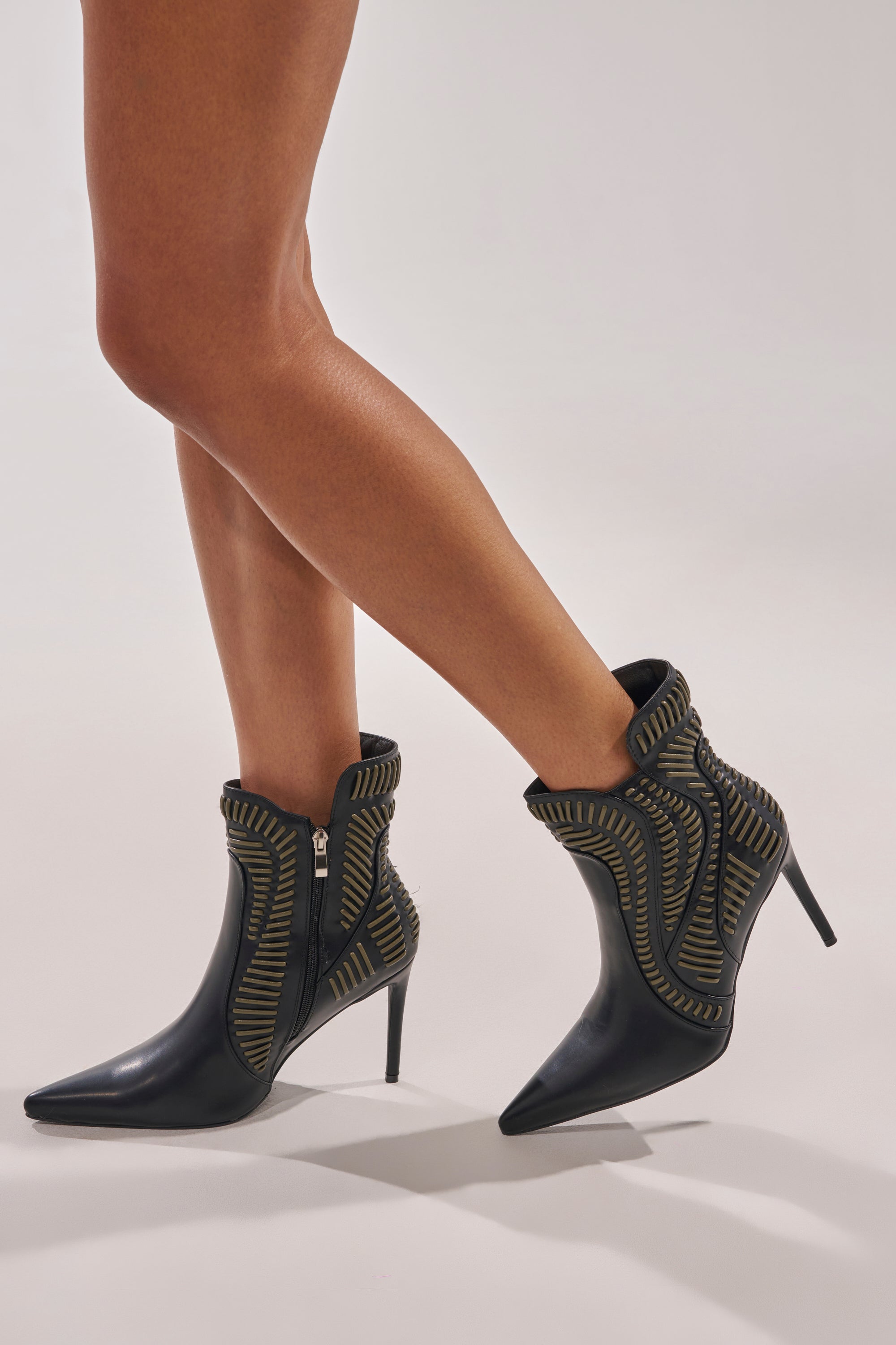 A person wears the AKIRA GOLD WEAVER BLACK AND OLIVE BOOTIE—black stiletto ankle boots with pointed toes, yellow stitching, side zip closure, and faux leather—while standing on a light-colored background. 2