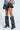 A person wears the AZALEA WANG WELLS BLACK EMBELLISHED BOOT, featuring knee-high black boots with a chunky heel and silver stud circular patterns. They pair it with a shiny black skirt and hold a zebra-patterned vegan leather handbag against a light grey background.