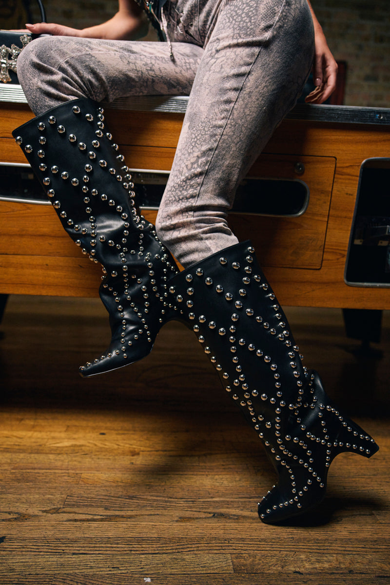 A person sits on the edge of a wooden table, one leg crossed over the other, wearing snakeskin-patterned pants and AZALEA WANG WELLS BLACK EMBELLISHED BOOTS adorned with silver studs.