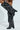 The AZALEA WANG WELLS BLACK EMBELLISHED BOOT is a striking vegan leather knee-high with silver studs, a low chunky heel, and pointed toe, worn by someone standing cross-legged against a white background.