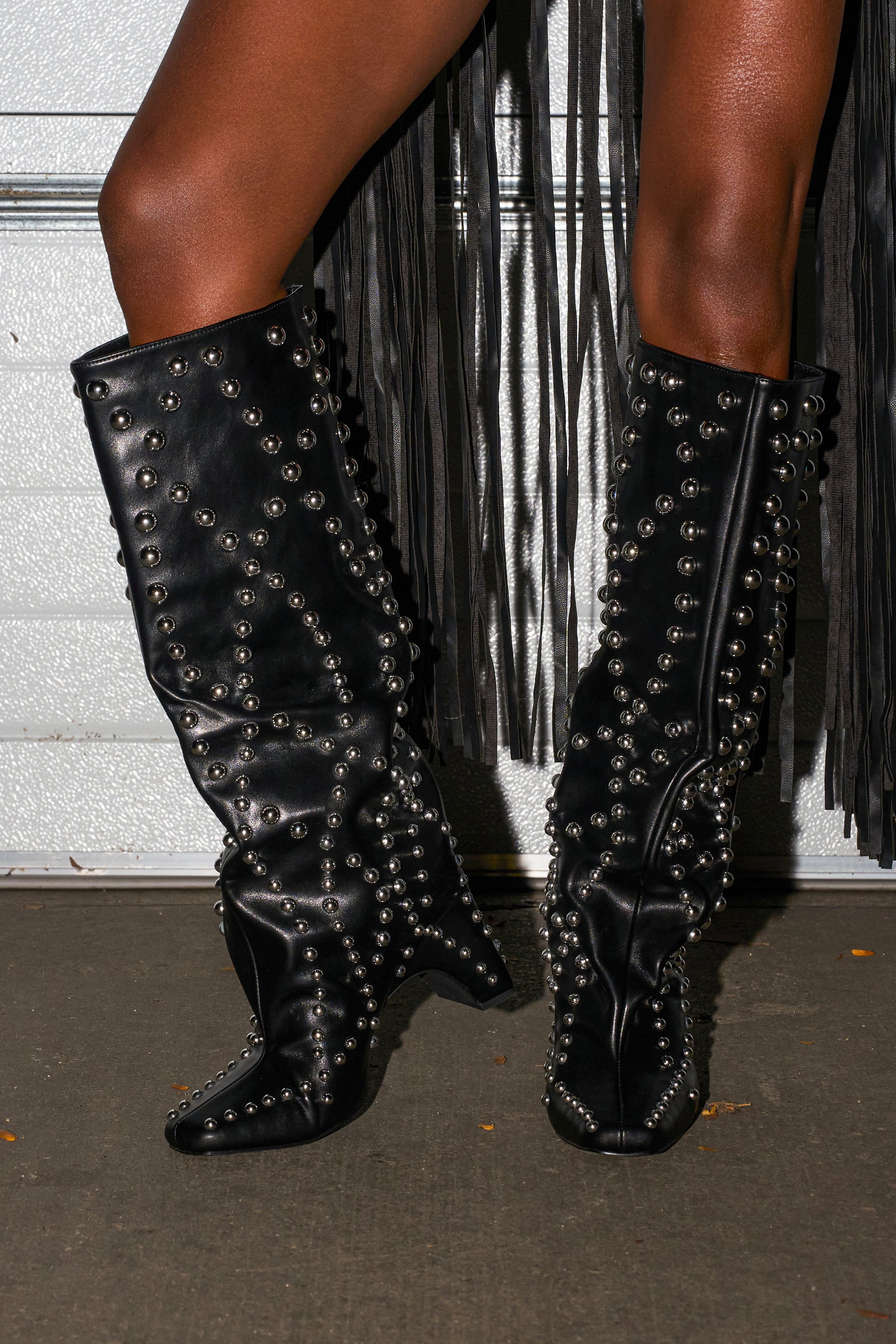 Someone stands on a concrete floor wearing the AZALEA WANG WELLS BLACK EMBELLISHED BOOT, which features a tall silhouette and chunky heel; long black fringe from a garment hangs nearby.