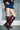 A person wearing a denim mini skirt, maroon top, and AZALEA WANG WELLS NEW BURGUNDY CROC BOOTS stands indoors in front of a glass block wall.