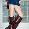 A person wearing a denim mini skirt, maroon top, and AZALEA WANG WELLS NEW BURGUNDY CROC BOOTS stands indoors in front of a glass block wall.