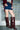 A person wearing a denim mini skirt, white crop top, burgundy blazer, and the AZALEA WANG WELLS NEW BURGUNDY CROC BOOT stands in front of a glass block wall.