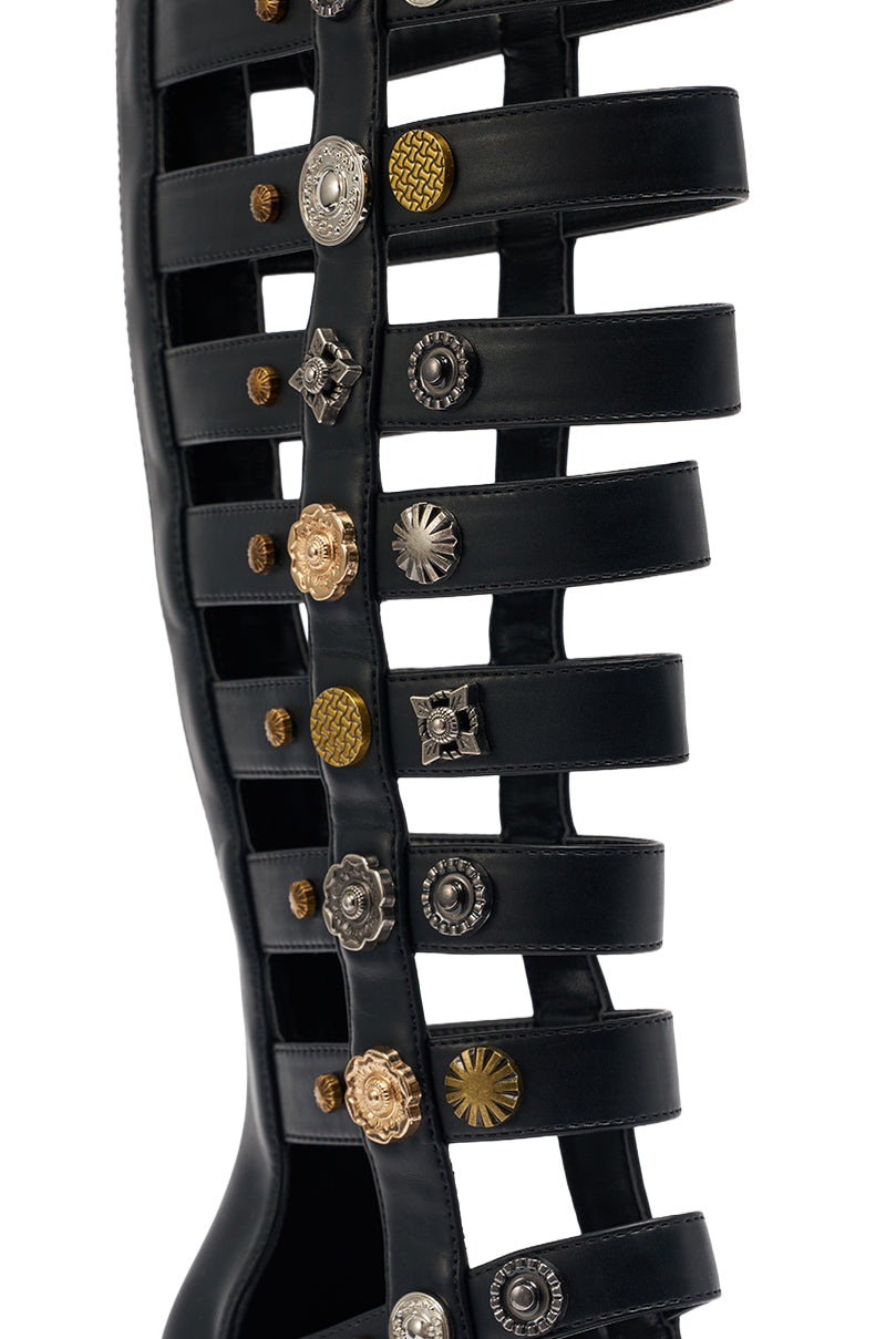 Close-up of the AZALWA WANG GOLD WENTWORTH GLADIATOR sandal, featuring black strappy material and two-toned gold and silver hardware embellishments with unique patterns, in an open toe knee-high design. 5