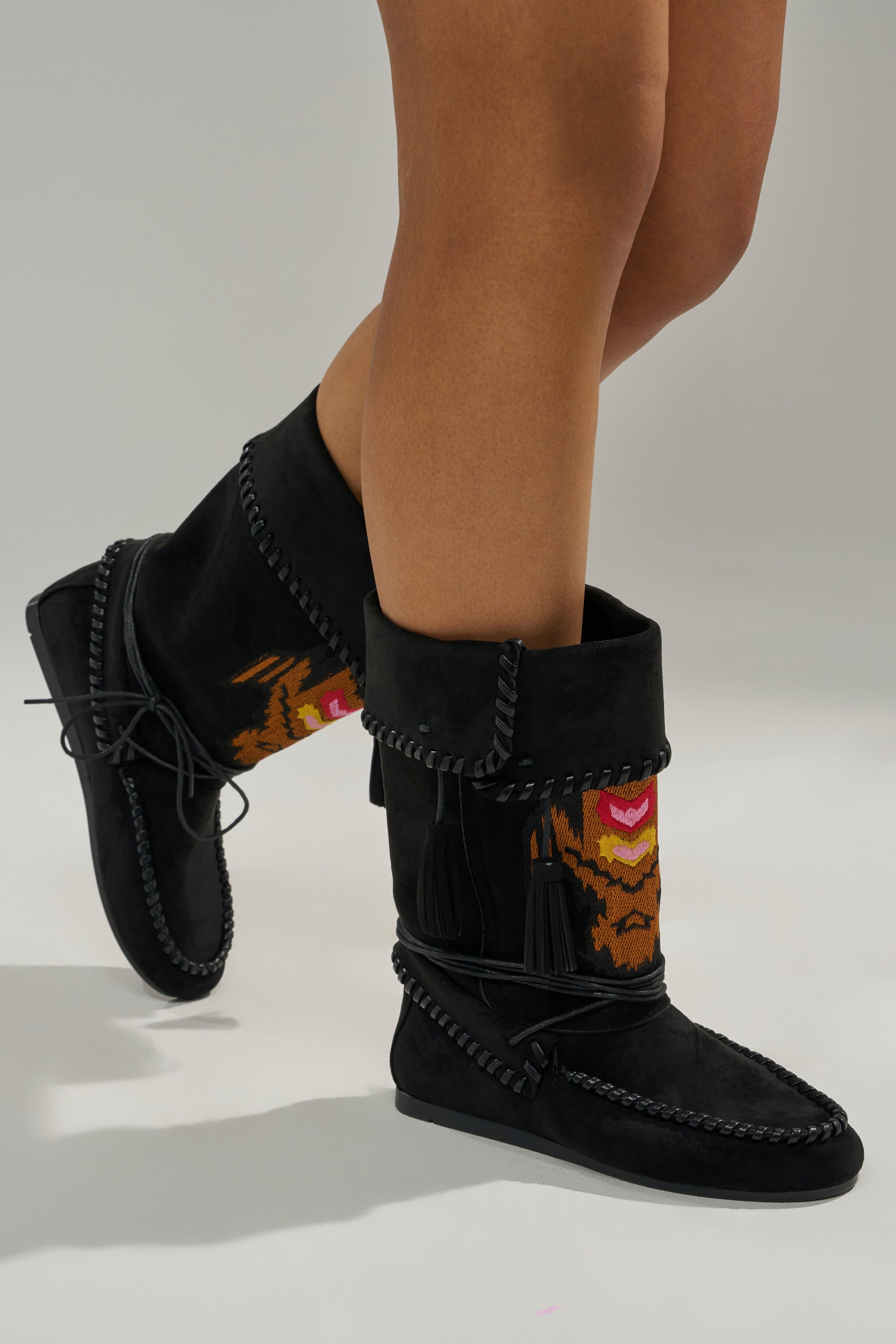 Someone wears the AZALEA WANG WERET BLACK FLAT BOOTIE, featuring black faux suede, tassels, and a woven orange, brown, and pink patterned shaft, while standing against a plain gray background.