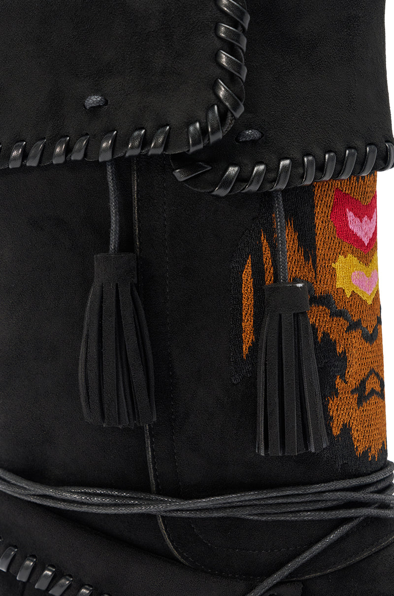 Close-up of the AZALEA WANG WERET BLACK FLAT BOOTIE in soft black suede, featuring leather whipstitch trim, black tassels, colorful red, yellow, and orange embroidery, and wrapped black cords on a chic mid-calf silhouette.