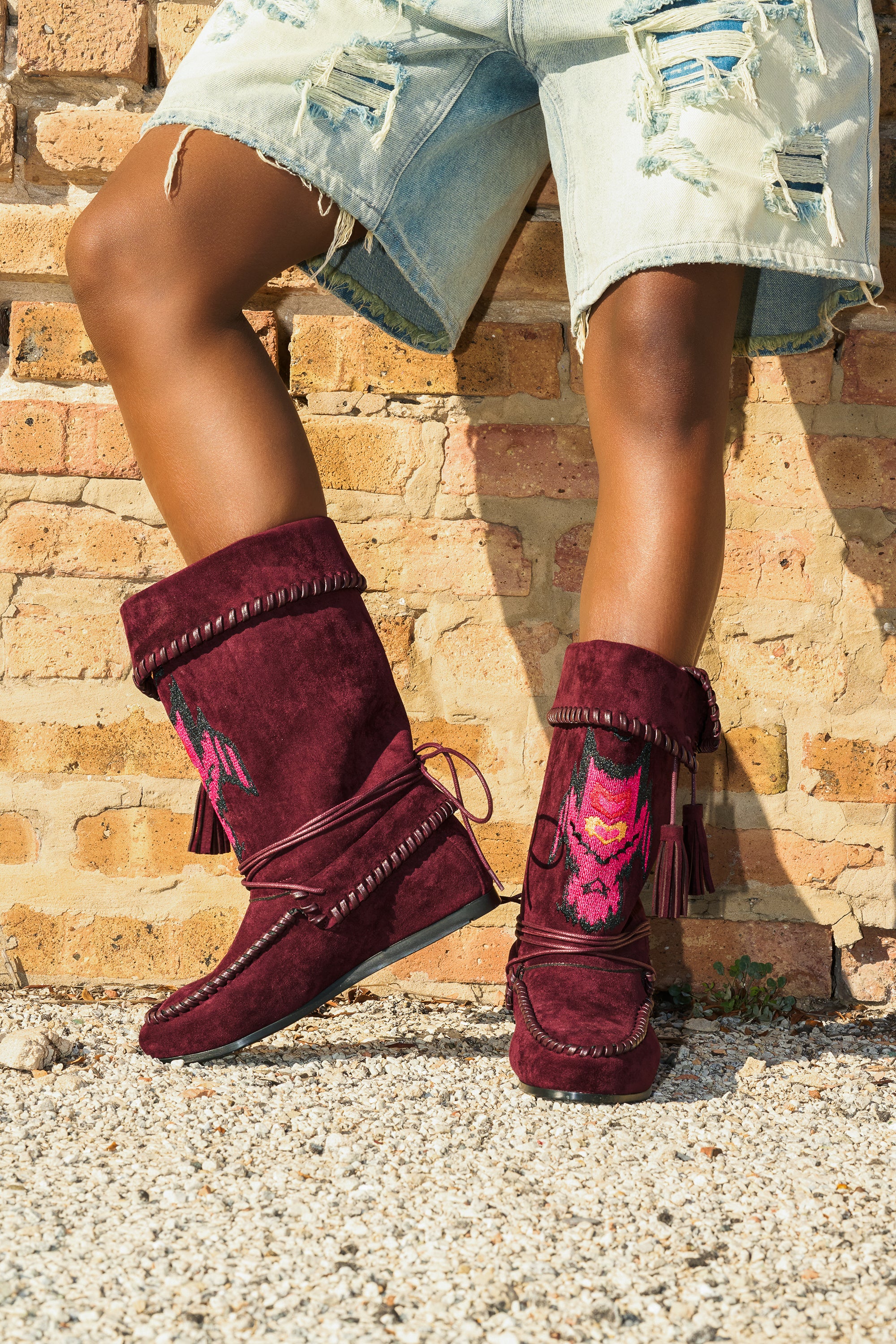 A person stands on gravel by a brick wall wearing distressed denim shorts and the AZALEA WANG WERET PLUM FLAT BOOTIE, featuring burgundy faux suede material with tassels and colorful embroidery.