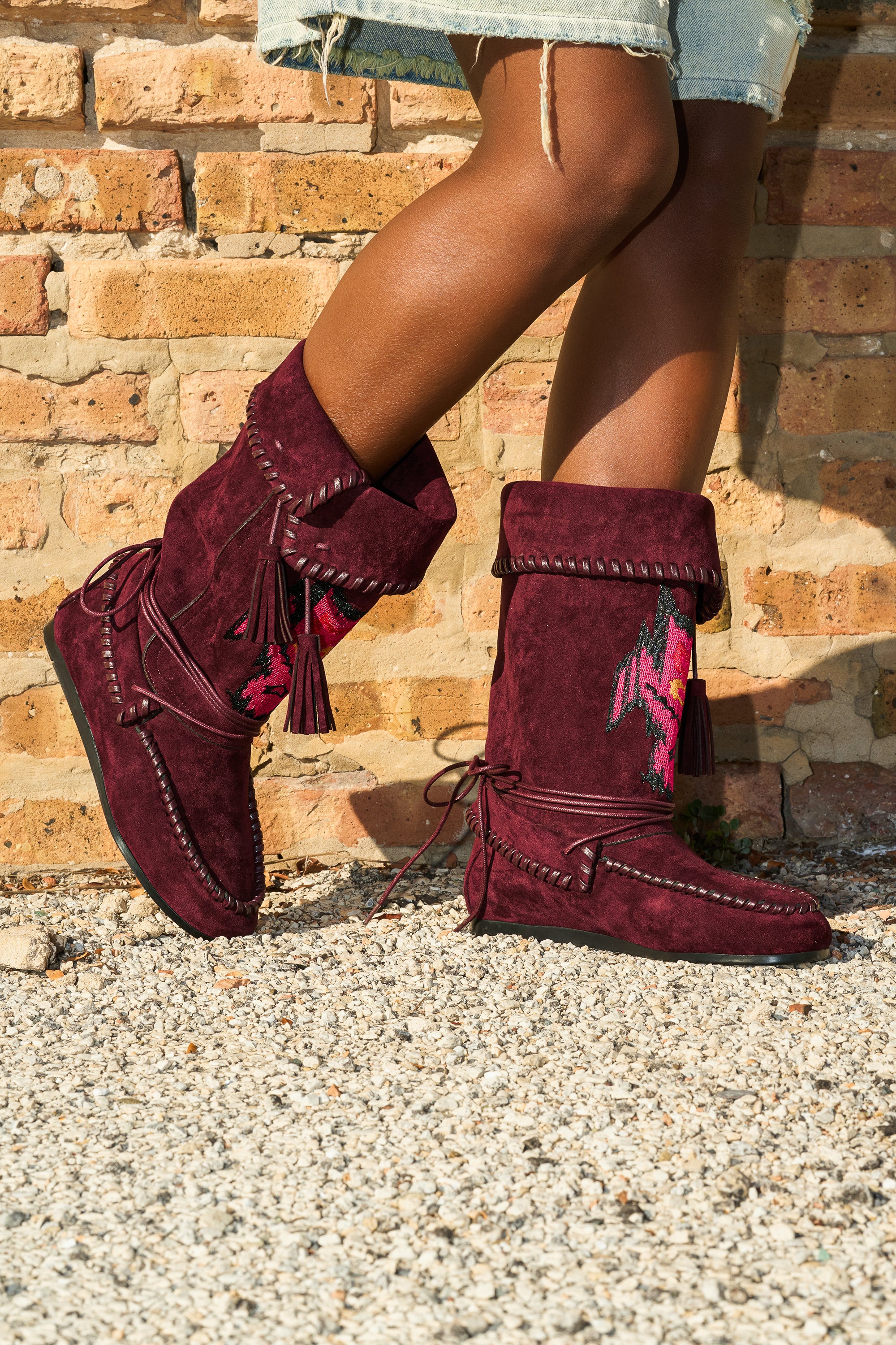 A person in knee-length denim shorts and AZALEA WANG WERET PLUM FLAT BOOTIE stands on gravel by a brick wall.