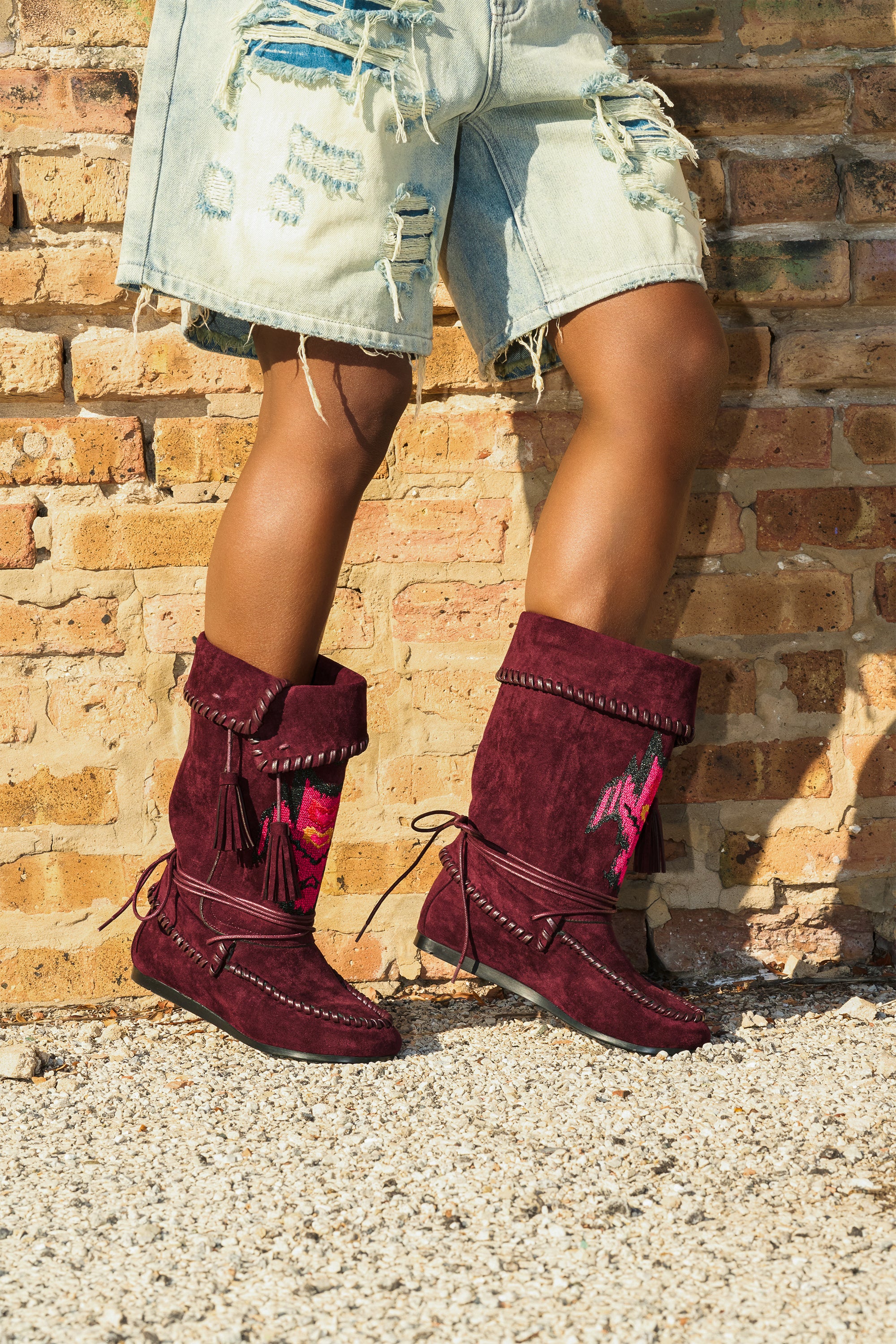 Someone stands on gravel before a rustic brick wall, wearing distressed denim shorts and the AZALEA WANG WERET PLUM FLAT BOOTIE, featuring fringe and tribal designs.