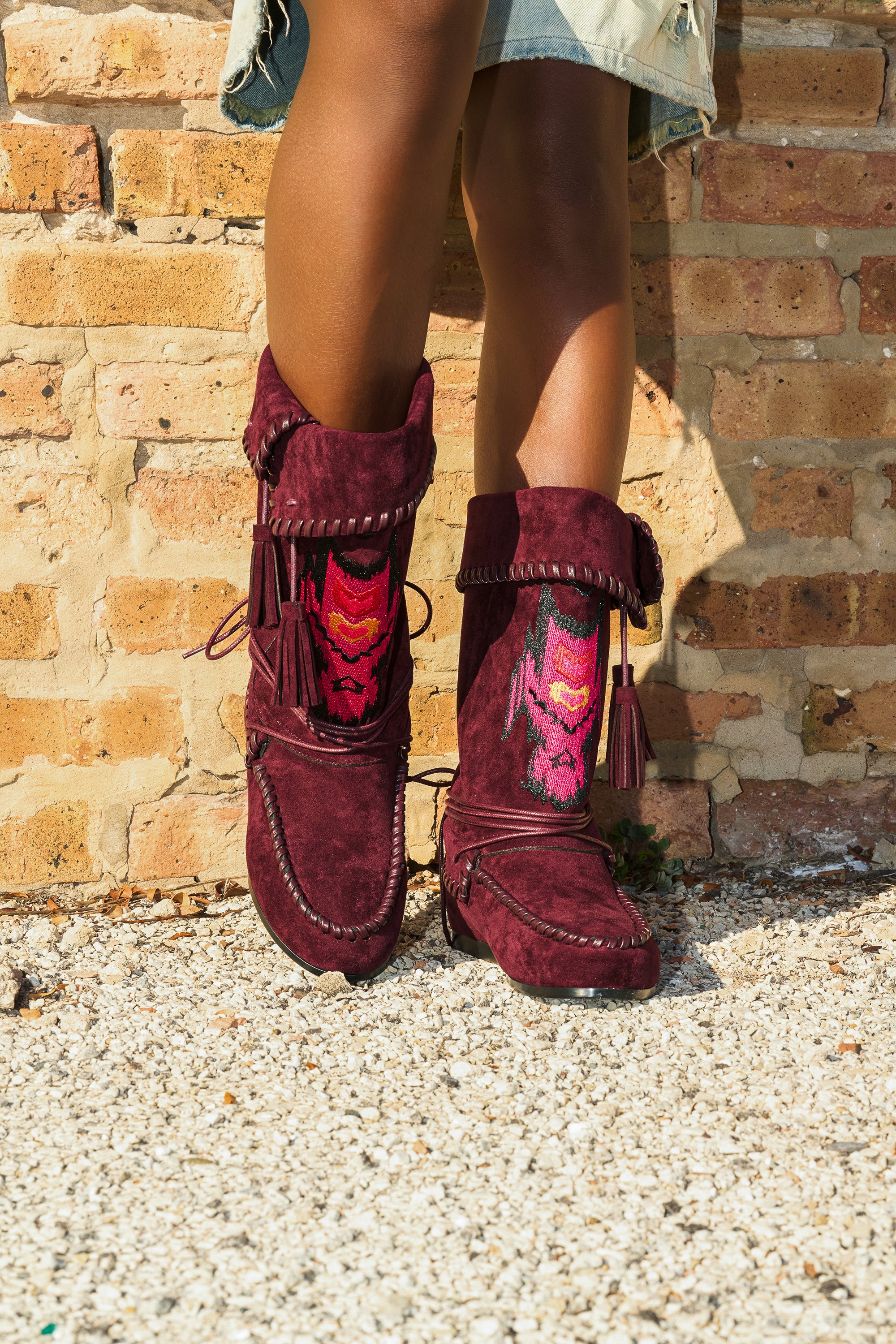 Wearing the AZALEA WANG WERET PLUM FLAT BOOTIE and colorful socks, a person stands on gravel by a brick wall, part of a light denim skirt visible.