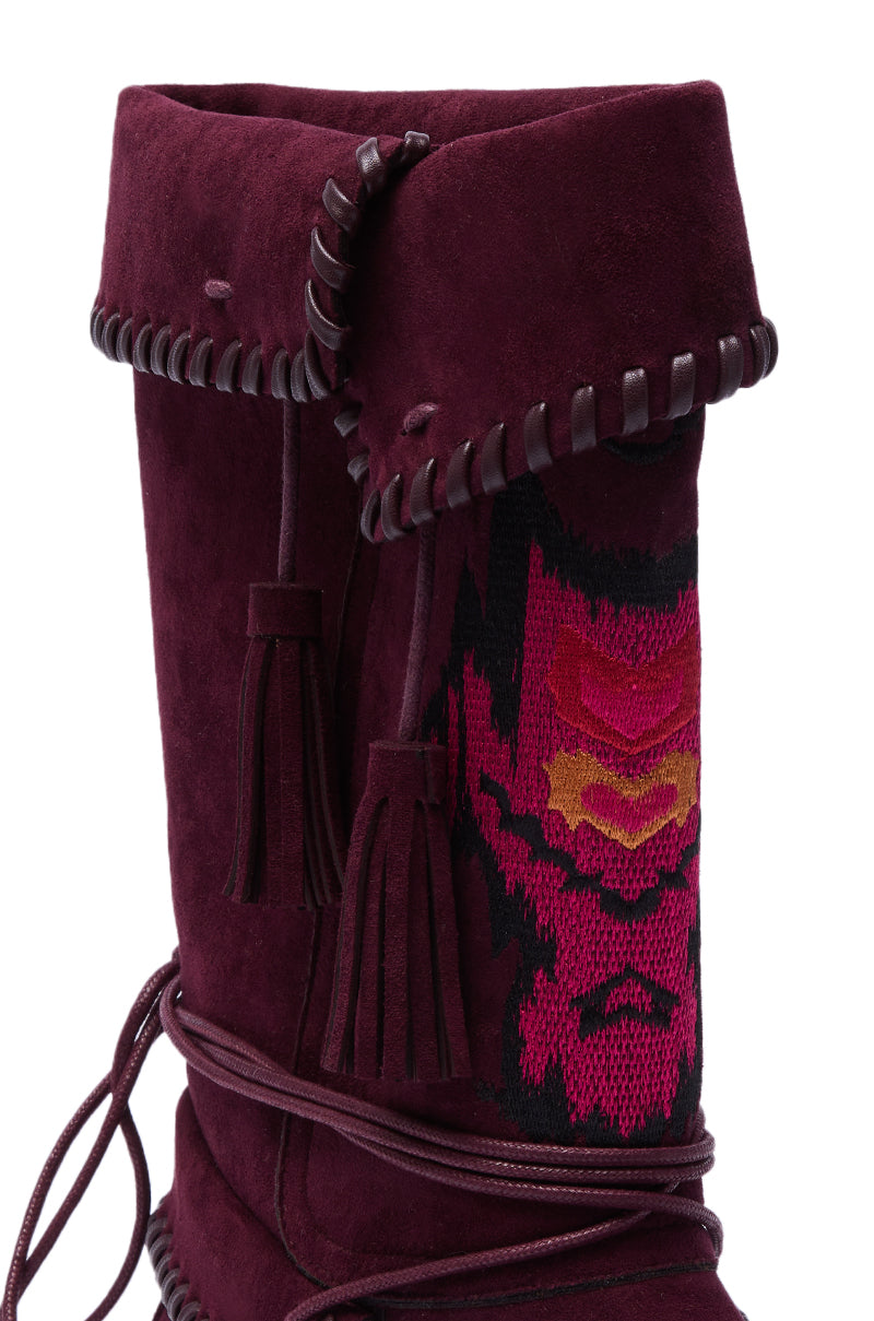 A close-up of the AZALEA WANG WERET PLUM FLAT BOOTIE, a tall burgundy faux suede boot with a folded cuff, leather whipstitch trim, tassel accents, and a decorative red, pink, and yellow woven side pattern. 6