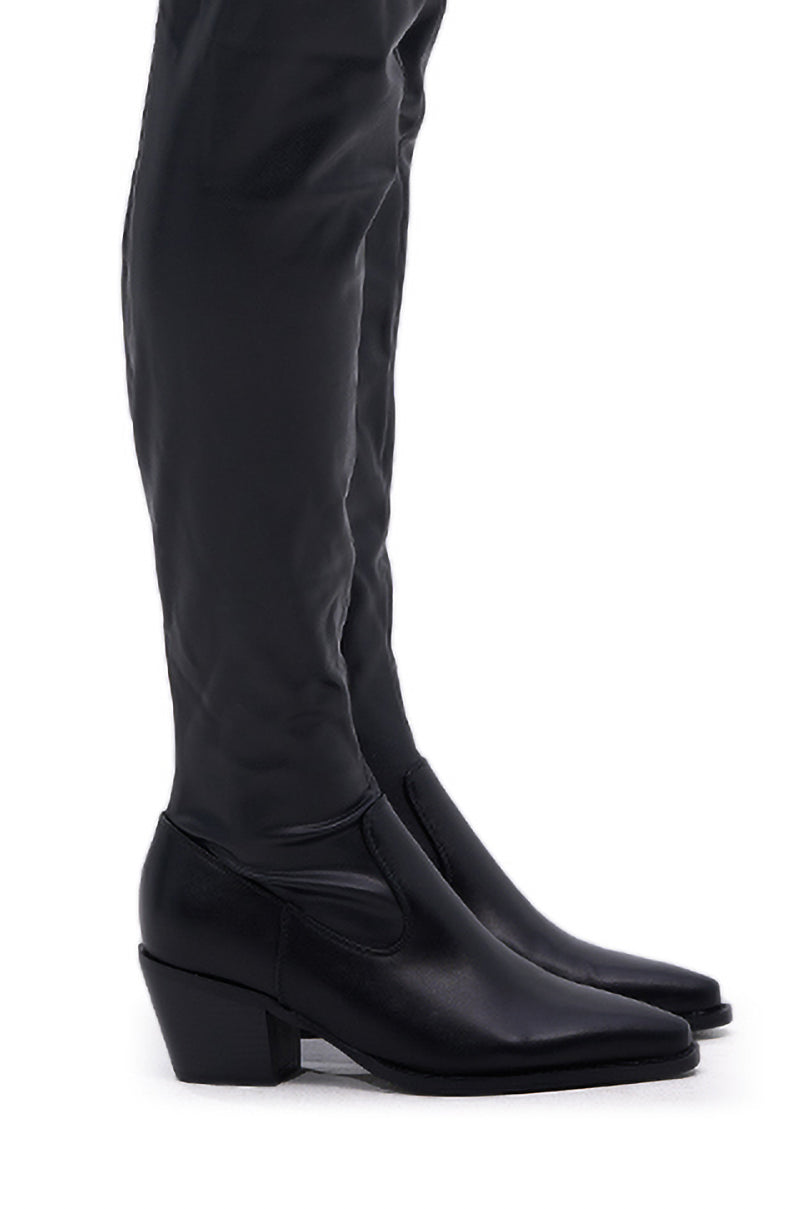 A close-up side view of a person wearing the AZALEA WANG WESTLANN BLACK PU CHAP BOOT, featuring sleek black faux leather with a pointed toe and fitted shaft, standing against a white background.